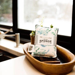 May include: A white and blue patterned sponge with the text "Naturally, "Naturally" SPONGE" on the front. The sponge is sitting in a wooden bowl.