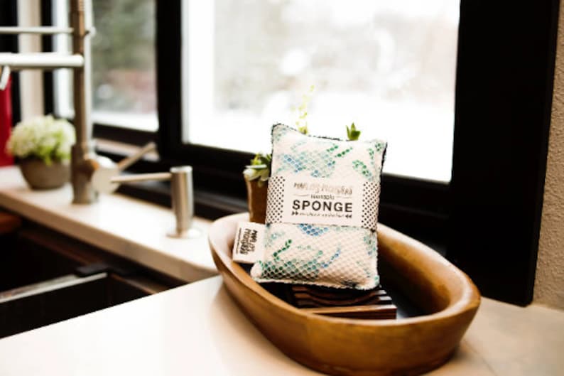 May include: A white and blue patterned sponge with the text "Naturally, "Naturally" SPONGE" on the front. The sponge is sitting in a wooden bowl.