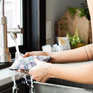 May include: A person rinsing a reusable produce bag under a kitchen faucet. The bag is made of a mesh material and has a colorful pattern. The faucet is silver and has a single handle.