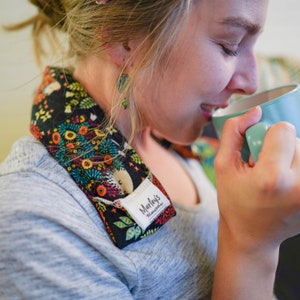 May include: A woman wearing a black and colorful floral neck wrap with the text "Marley's  Monogram" on it. She is holding a teal mug and drinking from it.