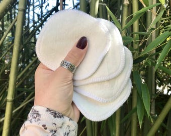 May include: A stack of six white reusable makeup remover pads with a stitched edge. The pads are held in a hand with a silver ring.
