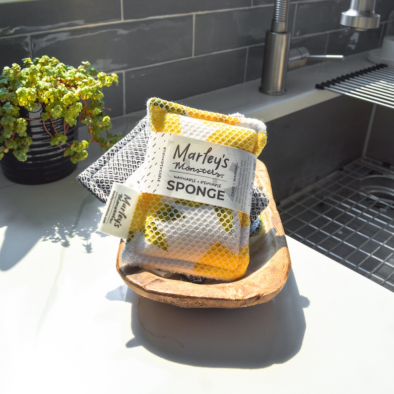 May include: Yellow and white dish sponge with a black and white label that reads "Marley's Monsters Washable + Reusable Sponge". The sponge is in a wooden bowl.