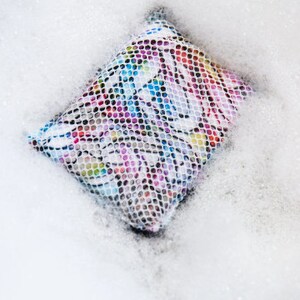 May include: A colorful laundry bag with a mesh design is submerged in soapy water. The bag is filled with bubbles and suds.
