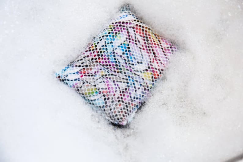 May include: A colorful laundry bag with a mesh design is submerged in soapy water. The bag is filled with bubbles and suds.