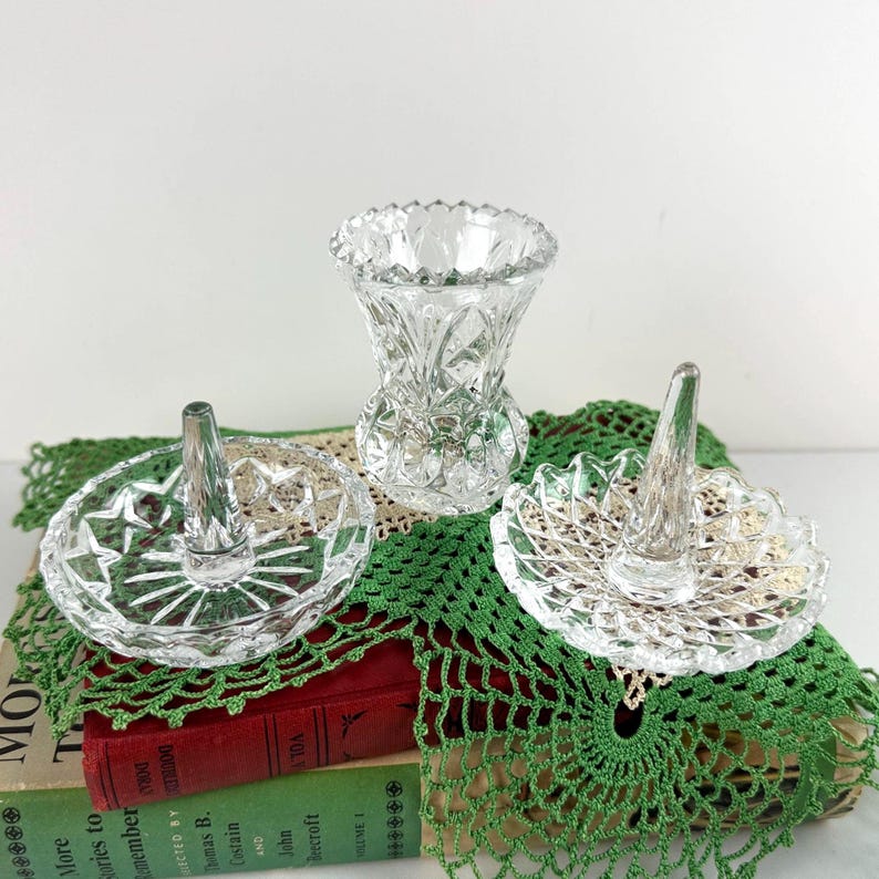 K&ouml;nnte beinhalten: Zwei Ringhalter aus klarem Glas mit runden, gewellten R&auml;ndern und einem zentralen konischen Stift, begleitet von einer kleinen Vase aus klarem Glas mit einem facettierten Diamantschliffmuster auf der gesamten Au&szlig;enfl&auml;che.