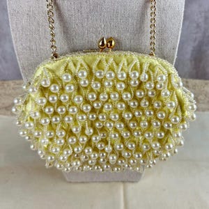 May include: A pale yellow satin handbag featuring an intricate overlay of white faux pearls arranged in a lattice pattern. The bag includes a gold-toned metal chain strap and a dual-knob clasp closure at the top, providing a secure fastening.