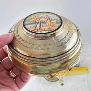 May include: A vintage brass trinket box with a decorative lid featuring a painted scene of two figures. The box has a round shape with a clear, decorative handle and a yellow comb is visible in the background.