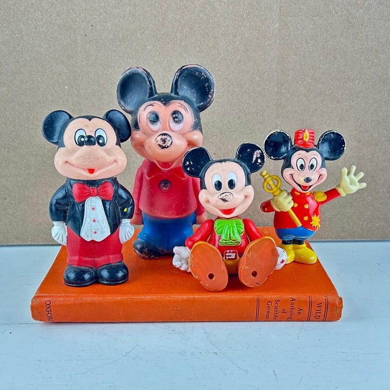 May include: Four vintage Mickey Mouse figurines in various poses and outfits, including a tuxedo, red shirt, and conductor's uniform. The figurines are displayed on a vintage orange book.
