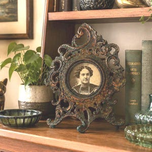 May include: A decorative tabletop picture frame with an ornate, dark metallic finish featuring intricate scrollwork and a circular center opening. The frame holds a black and white portrait of a person with dark hair wearing a high-collared garment.