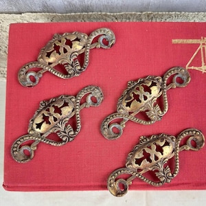 May include: Four matching brass drawer pulls featuring an ornate, openwork design with a central oval motif, intricate scrollwork, and beaded edges. Each piece includes two circular mounting holes at the ends for attachment to furniture surfaces.