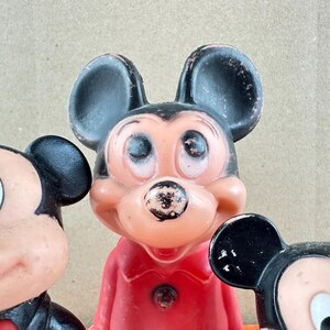 May include: Close-up of three vintage Mickey Mouse figurines. The figurines have black ears, a pink face, and a red outfit. The Mickey Mouse figurines are a classic Disney collectible.