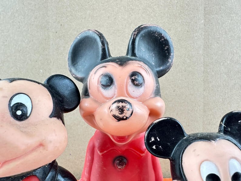 May include: Close-up of three vintage Mickey Mouse figurines. The figurines have black ears, a pink face, and a red outfit. The Mickey Mouse figurines are a classic Disney collectible.