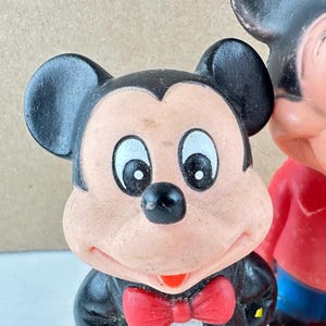 May include: Close-up of a vintage Mickey Mouse figurine. The figure has a black tuxedo, a red bow tie, and a smiling face with large white eyes. The ears are black and rounded. The figurine is a classic Disney collectible.