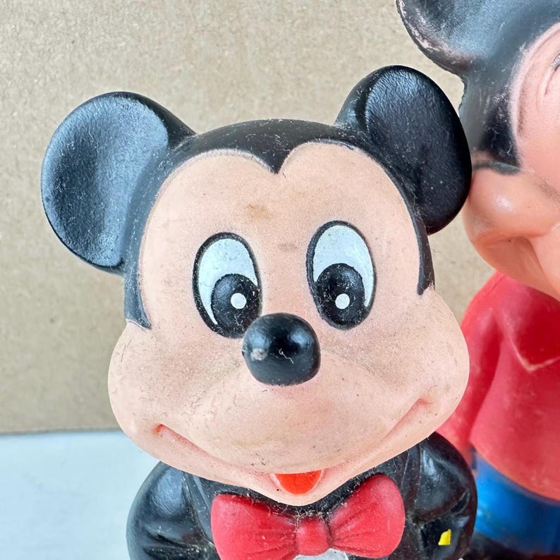 May include: Close-up of a vintage Mickey Mouse figurine. The figure has a black tuxedo, a red bow tie, and a smiling face with large white eyes. The ears are black and rounded. The figurine is a classic Disney collectible.