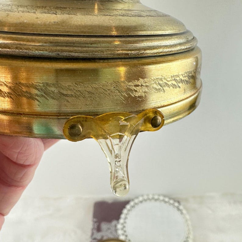 May include: Close-up of a vintage brass container with a decorative glass handle. The brass has a worn, antique finish. The glass handle is clear with a teardrop shape.