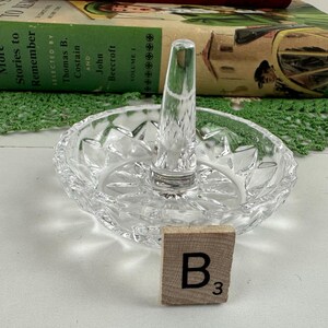 K&ouml;nnte beinhalten: Ein Ringhalter aus klarem Glas mit einer runden Basis, die ein facettiertes Sternmuster aufweist, und einem zentralen konischen Pfosten zur Aufbewahrung von Ringen. Ein kleines quadratisches Holzpl&auml;ttchen mit dem Buchstaben B und der Zahl 3 liegt davor.