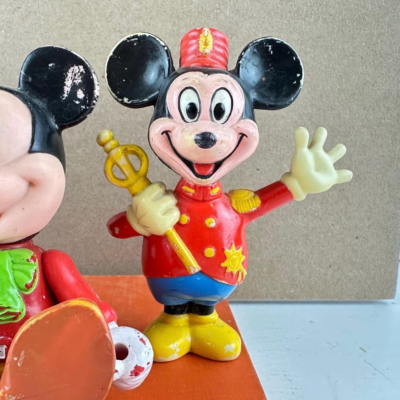 May include: Vintage Mickey Mouse figurine in a red band uniform with gold accents, blue pants, and yellow shoes. The figurine is holding a gold scepter and has a smiling expression. The black ears are visible.