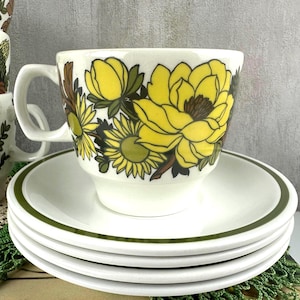 May include: A white ceramic teacup and saucer set featuring a bold floral pattern with large yellow blossoms, dark green leaves, and brown accents. The saucer has a simple dark green rim, and the cup has a sturdy handle and a tapered base.