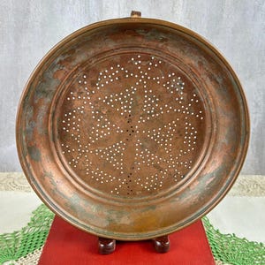 May include: A round copper colander featuring a wide rim and a central base with numerous small, evenly spaced drainage holes. The metal surface displays a weathered, oxidized patina with visible variations in color ranging from brown to green.