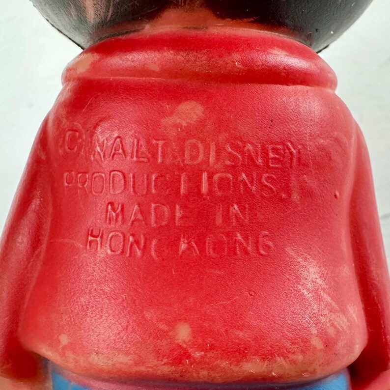May include: Close-up of a vintage toy figure with a red coat and black head. The text "&copy; WALT DISNEY PRODUCTIONS. MADE IN HONG KONG" is embossed on the back of the coat. The toy has blue and pink accents.