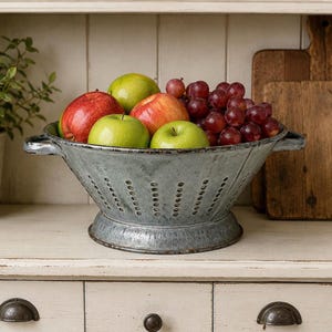 May include: A galvanized metal colander filled with fresh fruit, including red and green apples, and red grapes. The colander has a distressed finish and is sitting on a white surface.