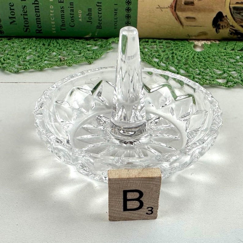 K&ouml;nnte beinhalten: Ein Ringhalter aus klarem Glas mit einer runden Schale, die ein Sternmuster aufweist, und einem zentralen konischen St&auml;nder. Ein kleines quadratisches Holzpl&auml;ttchen mit dem schwarzen Aufdruck B3 steht vor dem Ringhalter.