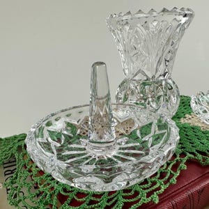 K&ouml;nnte beinhalten: Ein Ringhalter aus klarem Glas mit einer kreisf&ouml;rmigen Basis mit Sternmuster und einem zentralen konischen Pfosten. Hinter dem Ringhalter steht eine passende Vase aus klarem Glas mit facettiertem Design und einem gewellten oberen Rand.