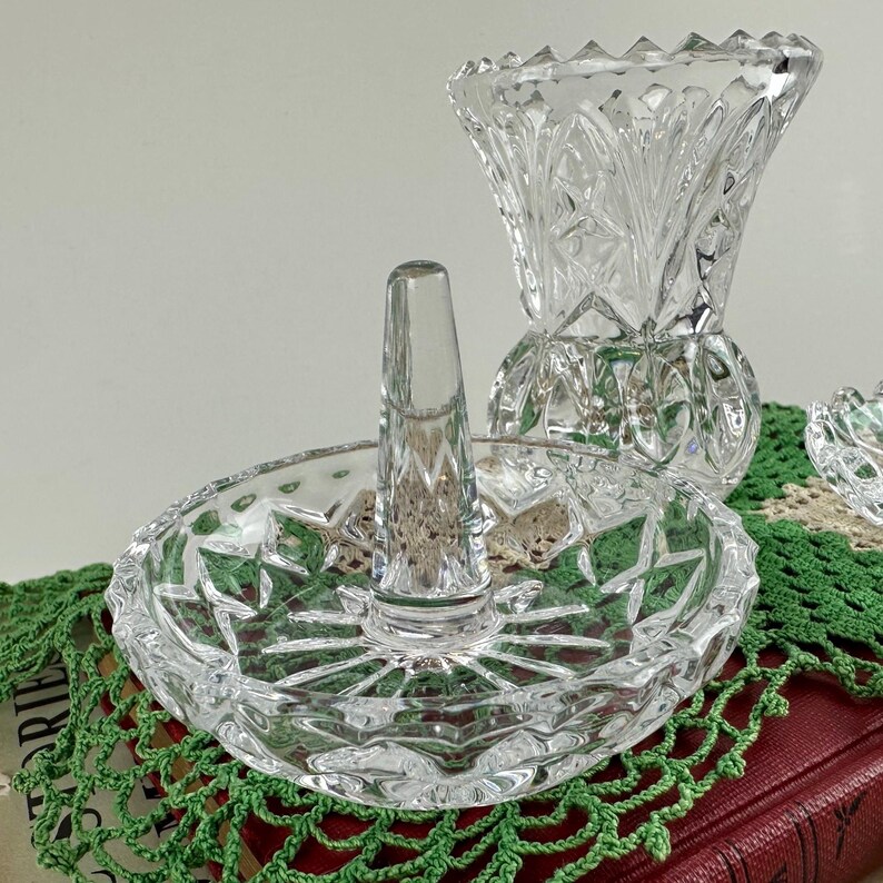 K&ouml;nnte beinhalten: Ein Ringhalter aus klarem Glas mit einer kreisf&ouml;rmigen Basis mit Sternmuster und einem zentralen konischen Pfosten. Hinter dem Ringhalter steht eine passende Vase aus klarem Glas mit facettiertem Design und einem gewellten oberen Rand.