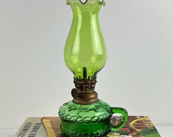 Puede incluir: Una lámpara de aceite de vidrio verde con una chimenea de borde ondulado y una base texturizada con asa. La lámpara incluye un quemador metálico con una perilla de ajuste circular, sobre una base con un patrón decorativo en relieve.