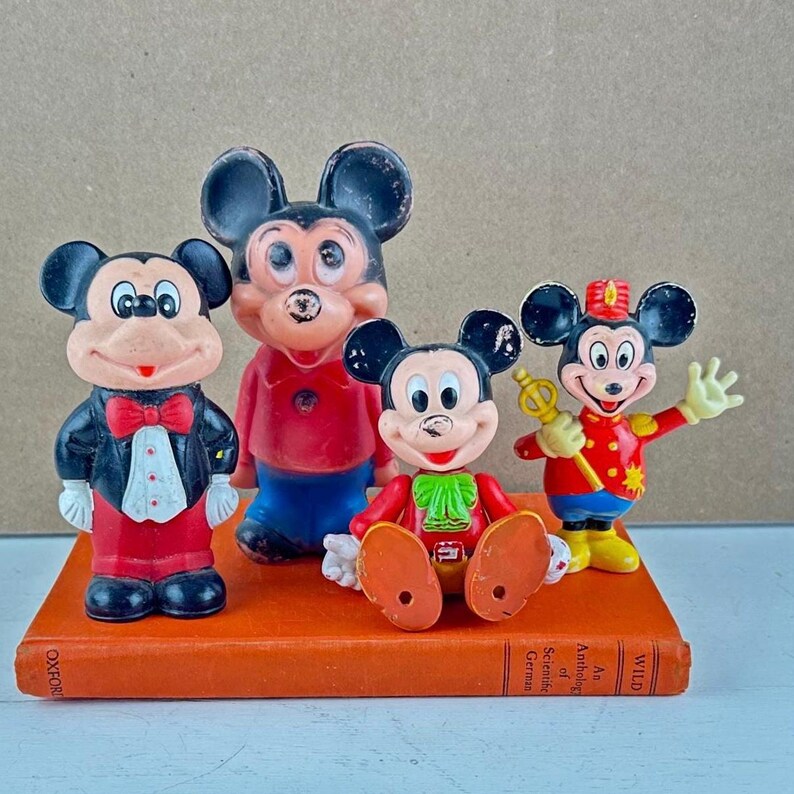 May include: Four vintage Mickey Mouse figurines in various poses, standing on an orange book. The figurines are in red, black, and white, with some wearing outfits. The book has text on the spine.