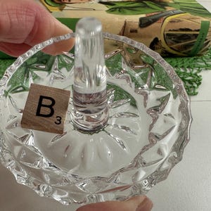 K&ouml;nnte beinhalten: Ein Ringhalter aus klarem Glas mit einer runden Schale, die einen facettierten Diamantschliff am &auml;u&szlig;eren Rand aufweist, sowie einem glatten, konischen Mittelpfosten f&uuml;r Ringe. Ein kleines quadratisches Holzpl&auml;ttchen mit dem Buchstaben B und der Zahl 3.