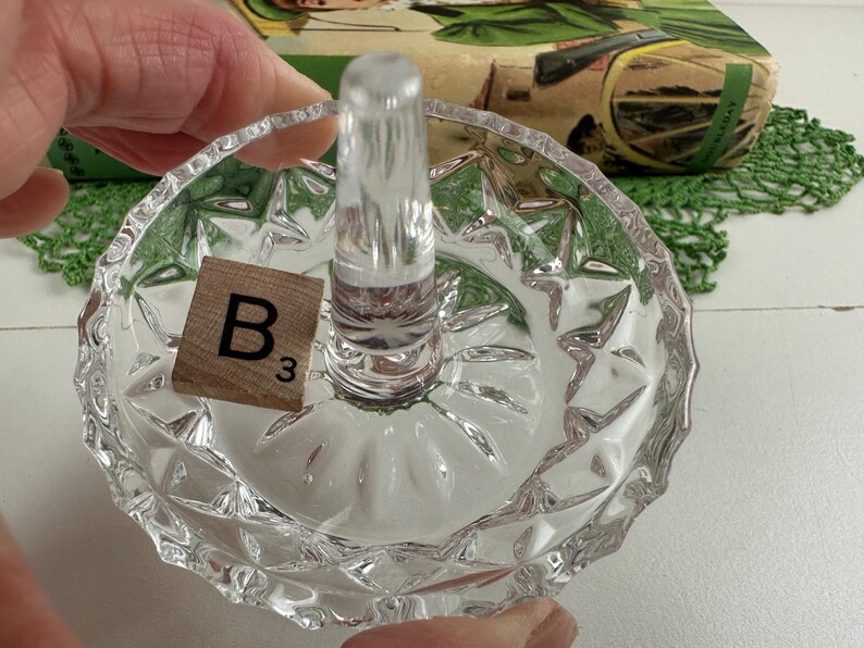 K&ouml;nnte beinhalten: Ein Ringhalter aus klarem Glas mit einer runden Schale, die einen facettierten Diamantschliff am &auml;u&szlig;eren Rand aufweist, sowie einem glatten, konischen Mittelpfosten f&uuml;r Ringe. Ein kleines quadratisches Holzpl&auml;ttchen mit dem Buchstaben B und der Zahl 3.