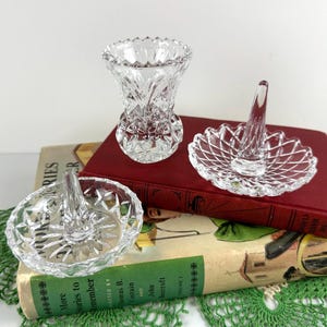 K&ouml;nnte beinhalten: Zwei klare Glas-Ringhalter mit gewelltem Rand und einem zentralen Kegel, begleitet von einer kleinen klaren Glasvase mit facettiertem Diamantschliff-Muster. Die Gegenst&auml;nde bestehen aus Pressglas und weisen eine reflektierende, transparente Oberfl&auml;che auf.