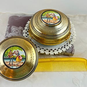 May include: Two antique gold-toned compacts and a yellow comb. The compacts feature a painted scene of a man playing a guitar and a woman dancing. The compacts are round and sit on a patterned fabric.