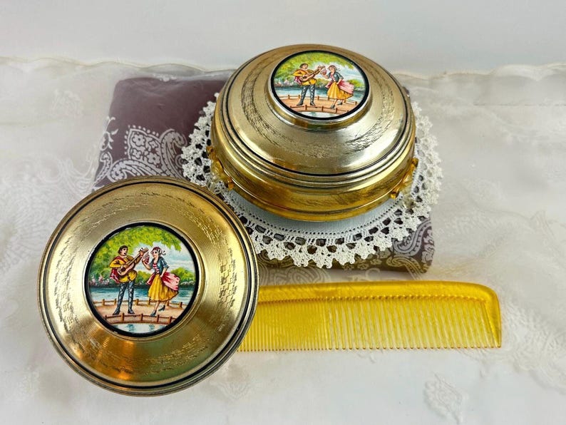 May include: Two antique gold-toned compacts and a yellow comb. The compacts feature a painted scene of a man playing a guitar and a woman dancing. The compacts are round and sit on a patterned fabric.