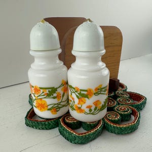 May include: A vintage salt and pepper shaker set with white ceramic bottles featuring a floral design in orange and yellow. The set includes a wooden holder and sits on a decorative green and red woven base.