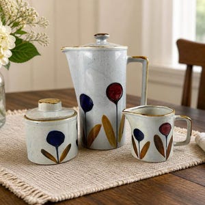 May include: A ceramic tea set with a light blue speckled finish, featuring a teapot, creamer, and sugar bowl. Each piece is adorned with a floral design in blue, red, and brown. The teapot has a tall, rectangular handle and a lid with a small knob.