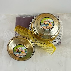 May include: Two gold-toned vintage powder compacts with a painted scene of musicians on the lids. A yellow comb rests between the compacts. The compacts sit on a purple pillow with a white lace trim.