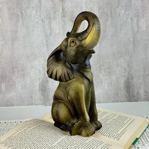 May include: A decorative elephant figurine crafted from a translucent, olive-green material with darker brown shading. The elephant is depicted in a seated position with its trunk curled upward, showcasing detailed textures on the ears and skin.