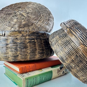 May include: Three woven baskets in varying sizes and shapes. The largest is round with a lid, the middle one is a bowl shape, and the third is a rectangular shape with a handle. The baskets are brown and tan, and sit on top of two books.