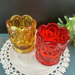 May include: Two small glass candle holders with a textured, starburst pattern. One is amber and the other is red.