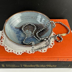May include: A gray pearl necklace draped over a blue and brown ceramic bowl. The bowl sits on a white lace doily, placed on top of an orange book. The book below is black with the title "Tender Is the Night".