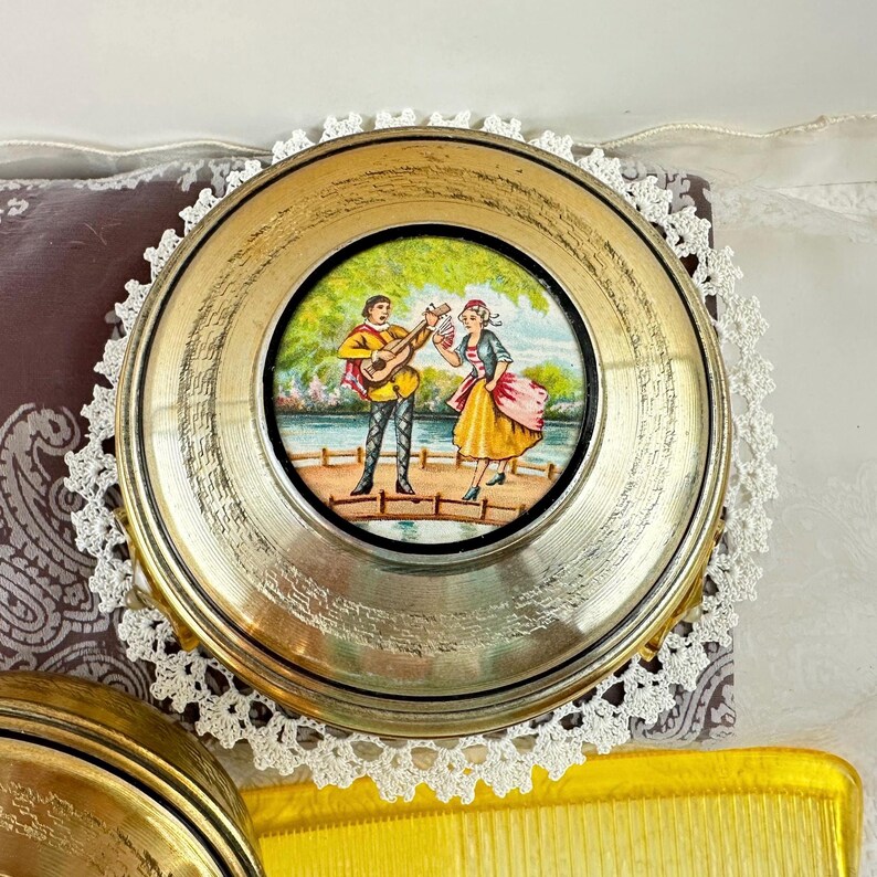 May include: A round, gold-toned compact mirror with a detailed painting of a man playing a guitar and a woman singing. The mirror is set on a white lace doily. A yellow comb is visible in the bottom right corner.