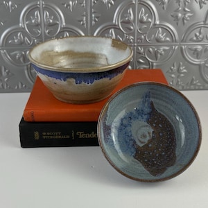 May include: Two handmade ceramic bowls. One bowl is tan with a blue rim, and the other is blue and brown. The bowls are sitting on top of two books. The background is a silver, patterned wall.