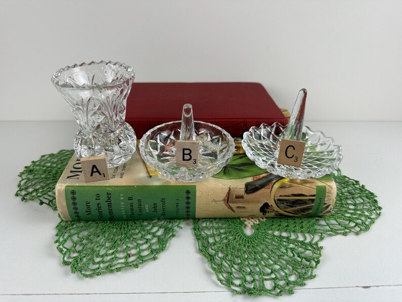 K&ouml;nnte beinhalten: Drei klare geschliffene Glasobjekte, beschriftet mit Holzpl&auml;ttchen: eine kleine Vase mit der Markierung A, eine Ringablage mit einem zentralen Kegel mit der Markierung B und eine zweite Ringablage mit einem zentralen Kegel mit der Markierung C.