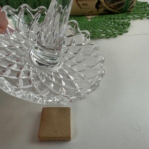 K&ouml;nnte beinhalten: Ein Ringhalter aus klarem Glas mit einem runden Fu&szlig; im Rautenmuster und einem zentralen, sich verj&uuml;ngenden St&auml;nder. Das Glas hat eine facettierte Textur, die das Licht reflektiert, und der Rand des Fu&szlig;es ist mit einem Wellenmuster versehen.