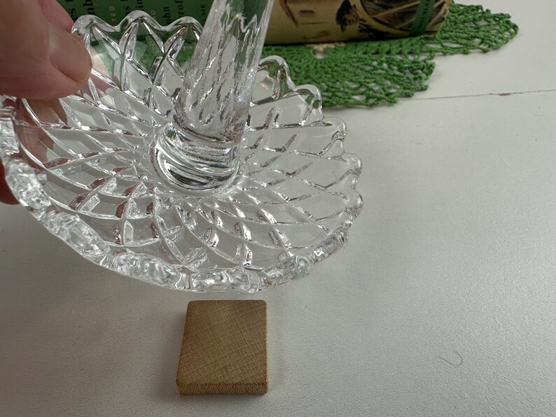 K&ouml;nnte beinhalten: Ein Ringhalter aus klarem Glas mit einem runden Fu&szlig; im Rautenmuster und einem zentralen, sich verj&uuml;ngenden St&auml;nder. Das Glas hat eine facettierte Textur, die das Licht reflektiert, und der Rand des Fu&szlig;es ist mit einem Wellenmuster versehen.