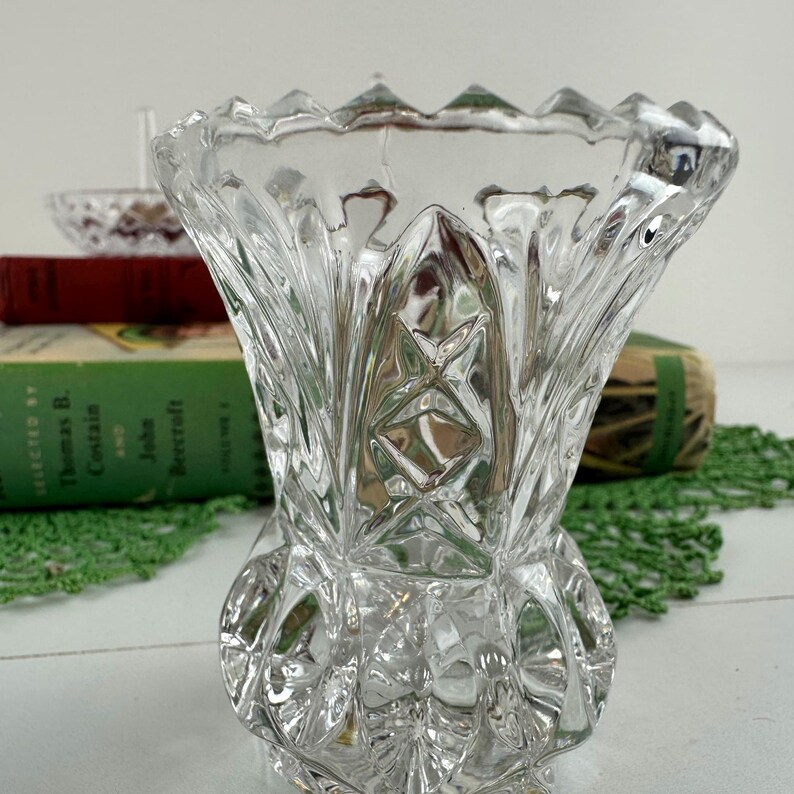 K&ouml;nnte beinhalten: Eine klare Glasvase mit einem facettierten, geometrischen Schliffmuster und einem gezackten, wellenf&ouml;rmigen Rand. Der K&ouml;rper der Vase zeigt ein sich wiederholendes Rauten- und Rippendesign, das eine strukturierte Oberfl&auml;che erzeugt, die das Licht reflektiert.
