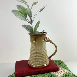 May include: A ceramic mug with a flared rim and a curved handle, featuring a two-tone glaze with a dark brown top and a textured, light tan base. The mug contains a green leafy plant stem with soft, matte-finished, oval-shaped foliage.