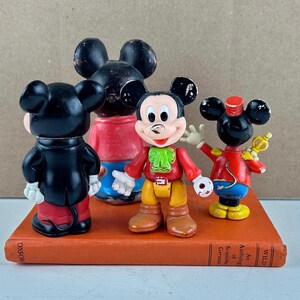 May include: Four vintage Mickey Mouse figurines in various poses, standing on a vintage book. The figures are in red, black, yellow, and white. One figure is waving, and another is dressed as a band leader.